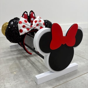 May include: A white, black, and red Minnie Mouse ear headband display stand. The stand holds several headbands, including a gold sequin ear, a black sequin ear, and a red and white polka dot bow.