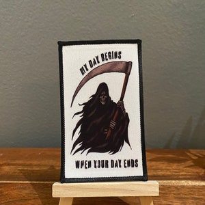 Op de afbeelding: Een rechthoekige patch met een zwarte rand, met een illustratie van de Grim Reaper met een zeis. De tekst luidt "MY DAY BEGINS" boven de figuur en "WHEN YOUR DAY ENDS" eronder. De patch wordt getoond op een klein houten ezel.