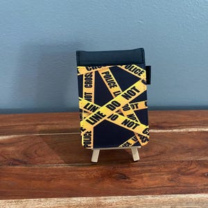 Op de afbeelding: Een zwart-gele telefoonhoes met een politie-afzetlint ontwerp. De hoes heeft de woorden "POLICE LINE DO NOT CROSS" in witte tekst op gele tape. De hoes wordt gepresenteerd op een klein houten ezel.