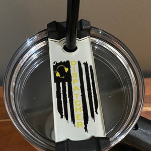 May include: A clear tumbler with a black lid and a black straw. A white tag with a black and yellow American flag design is attached to the lid. The tag says "Dispatcher" in yellow letters.