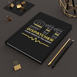 May include: Black journal with the text "911 DISPATCHER CALM VOICE IN THE DARK" in yellow and white. The cover design includes a thin yellow line and a US flag pattern. Gold and black pencils and paper clips are also visible.