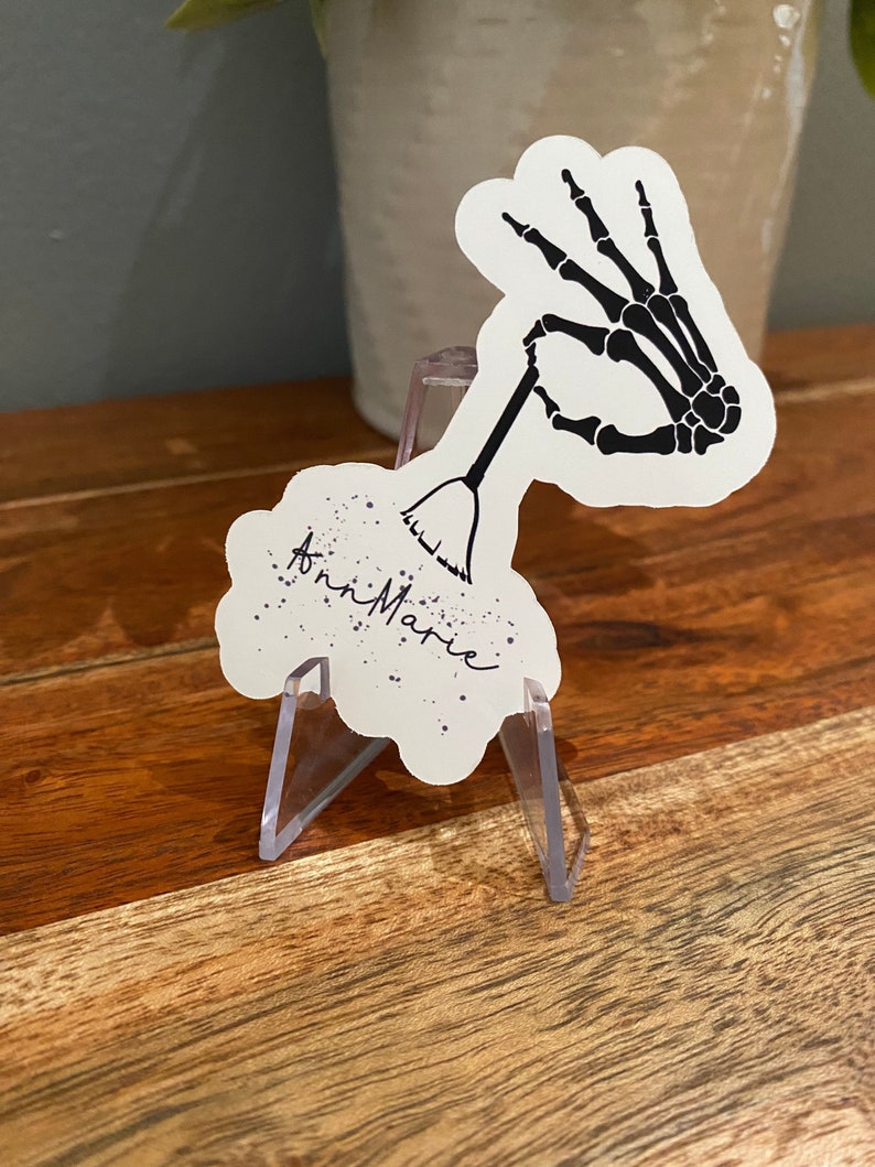 May include: A white sticker with a black skeleton hand holding a broom. The sticker has a cloud shape and the words "Ann Marie" are written in black ink.