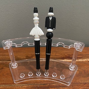 May include: Two black pens with decorative toppers. One topper is a white bride figurine with a white dress and a silver tiara. The other topper is a black groom figurine wearing a black tuxedo with a white shirt and a black bow tie.