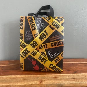 May include: Black tote bag with a crime scene theme. The bag features yellow "DO NOT CROSS" tape, a fingerprint, a body outline, a bullet, a ruler, and blood spatter graphics. The bag has a black handle.