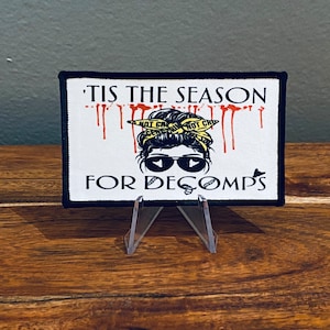May include: A white patch with black trim features a cartoon illustration of a woman with a yellow bandana and sunglasses. The text "'Tis the season for Decomps" is printed in black on the patch.