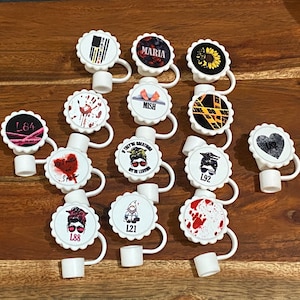 Op de afbeelding: Twaalf witte plastic flessenstoppers met verschillende ontwerpen. De ontwerpen omvatten een zonnebloem, een hart, een politielint, bloedspatten, een kabouter, een vrouw met een knot en een vrouw met een bandana. De stoppers hebben een gegolfde rand en een lus om op te hangen.