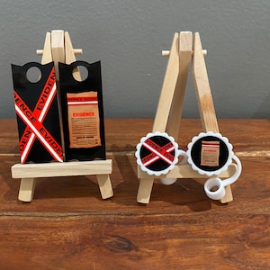 Op de afbeelding: Twee miniatuur ezelstandaards, een met een zwart en rood bewijslabel en de andere met twee ronde witte knoppen met zwart en rood bewijsontwerpen.