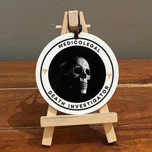 Peut inclure: Autocollant rond blanc avec un graphique de crâne noir et blanc et le texte "MEDICOLEGAL DEATH INVESTIGATOR". L'autocollant est sur un petit chevalet en bois.