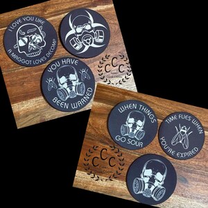 May include: Set of five black coasters with white designs. The coasters feature skulls, gas masks, flies, and text. The text on the coasters includes "I love you like a maggot loves decompose", "You have been warned", "When things go sour", "Time flies when you're expired".