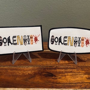 May include: Two white fabric patches with black borders, each featuring the word "FORENSICS" spelled out with various symbols representing forensic science, such as a skull, a measuring tape, a bullet, and a blood splatter. The patches are displayed on clear plastic stands.