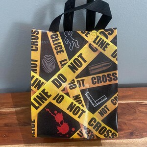 以下が含まれることがあります： 「POLICE LINE DO NOT CROSS」という文字が入った黄色い警察の立ち入り禁止テープ柄の黒いトートバッグ。指紋、弾丸、遺体のチョークアウトライン、定規の白いグラフィックアイコンと、赤い血痕のデザインが施されています。