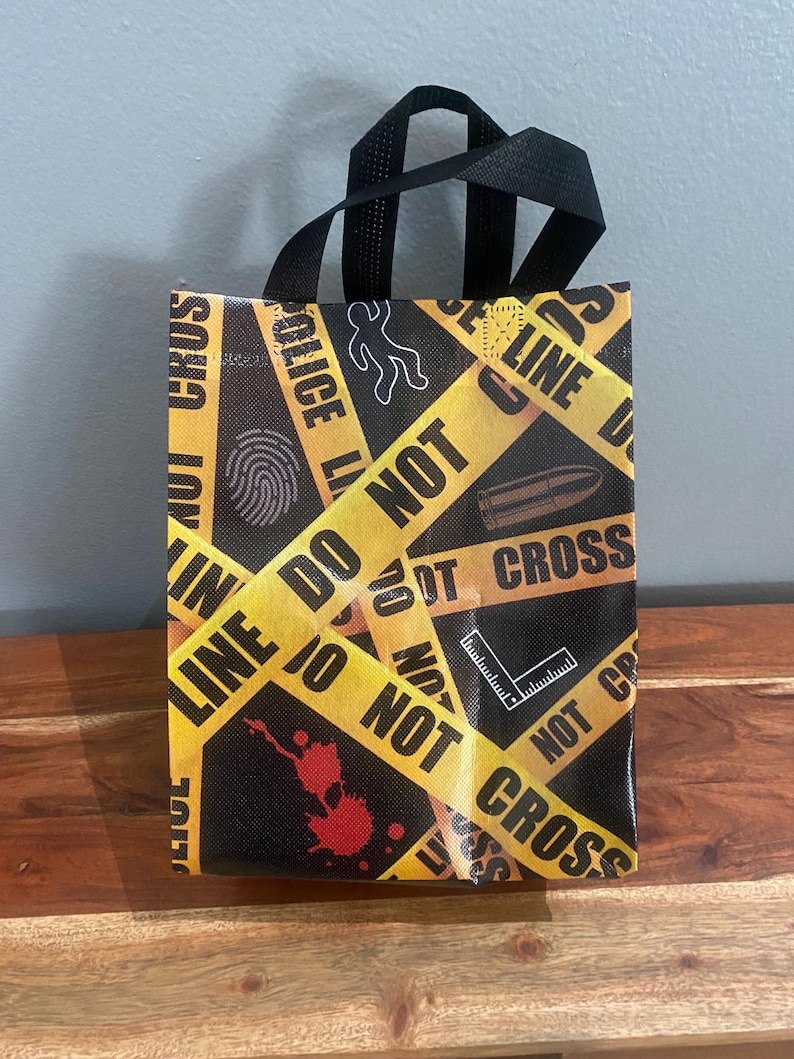 以下が含まれることがあります： 「POLICE LINE DO NOT CROSS」という文字が入った黄色い警察の立ち入り禁止テープ柄の黒いトートバッグ。指紋、弾丸、遺体のチョークアウトライン、定規の白いグラフィックアイコンと、赤い血痕のデザインが施されています。