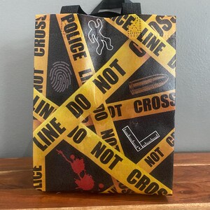 以下が含まれることがあります： POLICE LINE DO NOT CROSSという文字が入った黄色い警察の立ち入り禁止テープ柄の黒い再利用可能なトートバッグ。バッグには、チョークの輪郭、指紋、弾丸、直角定規の白いグラフィックアイコンと、赤い血の飛沫が描かれています。
