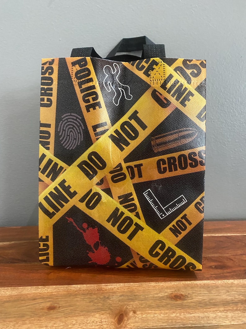 以下が含まれることがあります： POLICE LINE DO NOT CROSSという文字が入った黄色い警察の立ち入り禁止テープ柄の黒い再利用可能なトートバッグ。バッグには、チョークの輪郭、指紋、弾丸、直角定規の白いグラフィックアイコンと、赤い血の飛沫が描かれています。