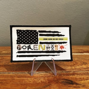 May include: A black and white American flag patch with the word "FORENSICS" spelled out in white letters with various crime scene related icons. The patch has a yellow "Crime Scene Do Not Cross" tape across the middle.