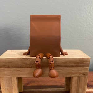 May include: A brown, rectangular figure with articulated legs and arms, seated on a small wooden bench. The figure's back is facing the viewer, with its arms resting on the bench. The legs are bent at the knees, and the feet are flat on the surface.