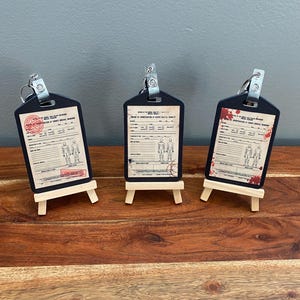 Puede incluir: Tres llaveros negros en forma de etiqueta con un diseño de gráfico médico vintage, cada uno montado en un pequeño caballete de madera. Los gráficos presentan texto, sellos e ilustraciones de figuras humanas. Un llavero tiene salpicaduras de sangre.