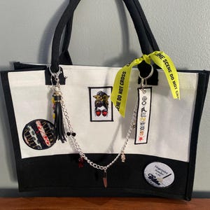 May include: A white and black tote bag featuring a central graphic of a person with a messy bun and sunglasses, a circular patch with red and black stripes, a rectangular patch with text, and a round patch reading Fingerprint powder is my glitter.