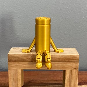 May include: A gold-colored, cylindrical figurine with arms and legs, sitting on a small wooden bench. The figurine has a detailed design on its head and is posed as if relaxing. The bench is made of light-colored wood.