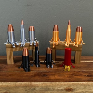 Op de afbeelding: Een verzameling antropomorfe kogelbeeldjes met koperkleurige punten. Drie zilveren en drie oranje figuren zitten op kleine houten bankjes, terwijl twee zwarte en één rode figuur op het oppervlak zitten, allemaal met dunne armen en benen.