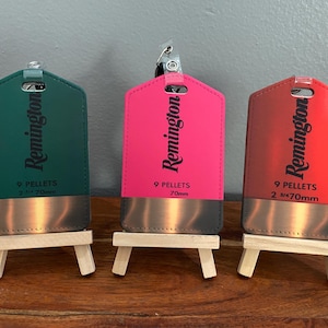 Może przedstawiać: Trzy kolorowe zawieszki marki Remington, zielona, różowa i czerwona, każda z metalową dolną częścią. Zawieszki są prezentowane na małych drewnianych sztalugach. Każda zawieszka ma napis "9 PELLETS 70mm".