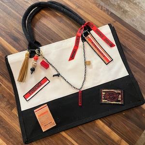 May include: A black and white canvas tote bag featuring black handles and multiple red and black patches with the word EVIDENCE printed on them. A black chain, a tan tassel, and a red tag labeled TOP SECRET are attached to the bag.