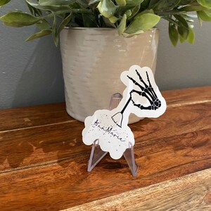 May include: A white sticker with a black skeleton hand holding a broom and the text "Anni Marie" in black cursive. The sticker is on a clear plastic stand.