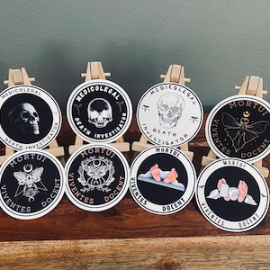 May include: Eight round stickers with black and white designs. The stickers feature a skull, a moth, and a pair of feet. The text on the stickers reads "Medicolegal Death Investigator" and "Mortui Viventes Docent".