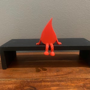 Puede incluir: Una figura caprichosa roja en forma de lágrima con brazos y piernas articuladas está posada sobre una pequeña mesa rectangular negra. Los brazos de la figura descansan sobre el borde de la mesa y sus piernas cuelgan.