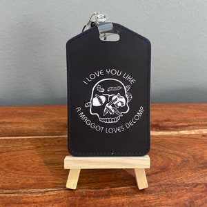 May include: Black silicone keychain with a white skull and maggots design. The text reads "I love you like a maggot loves decomp".