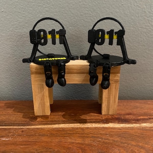 May include: Two black 3D-printed figurines of figures wearing headsets, with the number "911" in yellow. The figurines are seated on a small wooden bench, with the word "DISPATCHER" printed on the base.