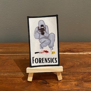 May include: A white patch with a cartoon illustration of a person in a white suit holding a camera, standing over a bloody knife and a yellow evidence marker. The text "FORENSICS" is printed in black below the illustration.