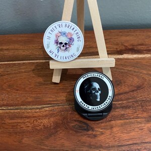 May include: Two phone grips with skull designs. The first grip is white with a skull surrounded by flowers and the text "If they're breathing we're leaving". The second grip is black with a skull and the text "Medico-legal Death Investigator".