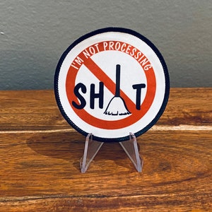 May include: A round, white patch with a red circle and a diagonal line through it. The text "I'm Not Processing Sh*t" is written around the circle. A black broom is inside the circle.