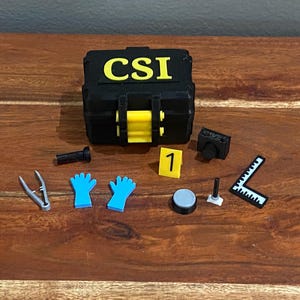 Puede incluir: Un kit negro de CSI con detalles amarillos y la palabra "CSI" en amarillo. El kit está rodeado de herramientas forenses en miniatura, incluyendo guantes azules, una cámara y una regla. El kit está sobre una superficie de madera.