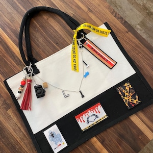 May include: A white and black canvas tote bag featuring a yellow crime scene tape ribbon, an evidence tag, and various charms including a camera, a red tassel, and letter charms. Three rectangular patches are attached to the lower black section.
