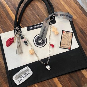 May include: A black and white canvas tote bag featuring the text LAST RESPONDER, a circular medical examiner patch, and a rectangular document patch. It is decorated with a silver chain, a grey tassel, and charms including a red heart, a tooth, and a torso.
