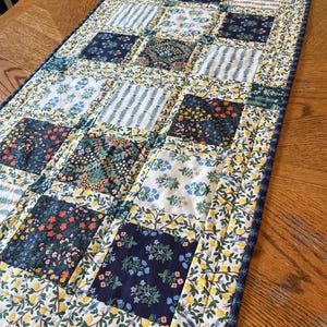 May include: A quilted table runner with a patchwork design. The runner features squares of floral and striped patterns in navy, white, yellow, and green. The edges are finished with a coordinating floral border. The runner is approximately 40 inches long.