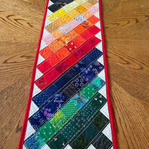 May include: A handmade, rectangular table runner with a vibrant rainbow design. The runner features a patchwork pattern of colorful fabric strips, including red, orange, yellow, green, and blue, bordered by white and a red edge. The runner is approximately 30 inches long.