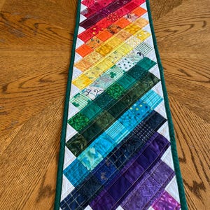 May include: A rectangular quilted table runner featuring a diagonal rainbow gradient pattern. The design consists of small, multi-patterned fabric squares in shades of red, orange, yellow, green, blue, and purple, bordered by a solid dark green trim.