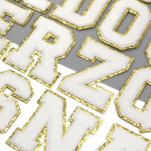 May include: White felt letters with gold glitter trim. The letters are in a varsity style font.