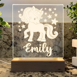 May include: A white acrylic night light with a unicorn and stars design. The name "Emily" is written below the unicorn. The light is on a wooden base.