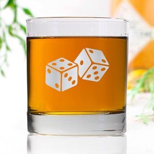 May include: A clear glass tumbler with a design of two dice etched on the side. The tumbler is filled with a light amber liquid.