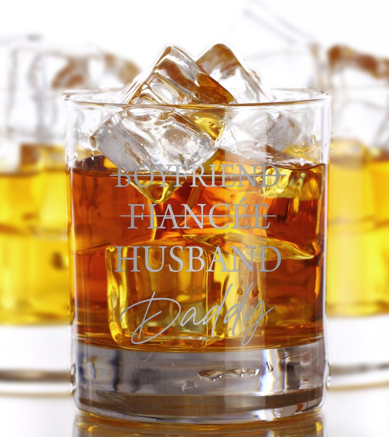 May include: A clear glass with a brown liquid and ice cubes. The glass has the words "Boyfriend - Fianc&eacute; - Husband Daddy" etched on the side.