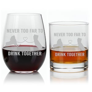 May include: Two clear drinking glasses, one stemless wine glass and one rocks glass, each etched with the words "NEVER TOO FAR TO DRINK TOGETHER". The wine glass is filled with red liquid, and the rocks glass contains amber liquid.