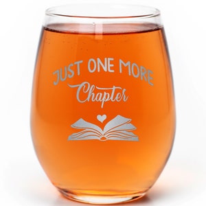 May include: A clear glass wine glass with a stem and a rounded bowl. The glass is filled with a light orange liquid. The glass has the text "Just One More Chapter" etched on the side, along with a stylized image of an open book with a heart in the center.