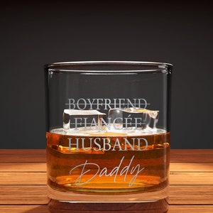 May include: A clear glass tumbler with the words "Boyfriend, Fianc&eacute;, Husband, Daddy" etched on the side. The glass is filled with amber liquid and ice cubes.