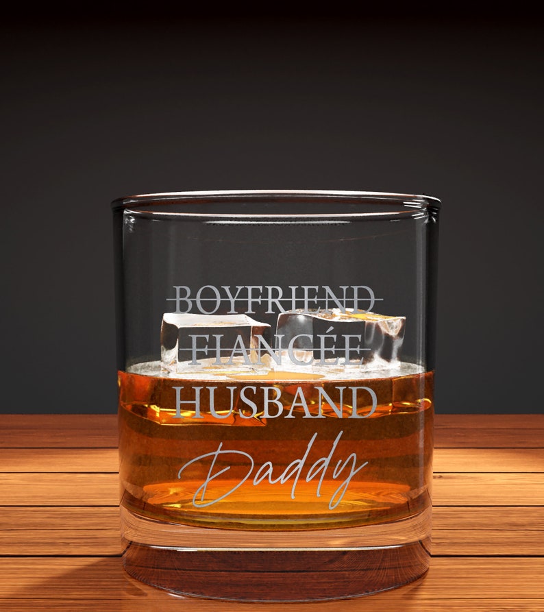 May include: A clear glass tumbler with the words "Boyfriend, Fianc&eacute;, Husband, Daddy" etched on the side. The glass is filled with amber liquid and ice cubes.