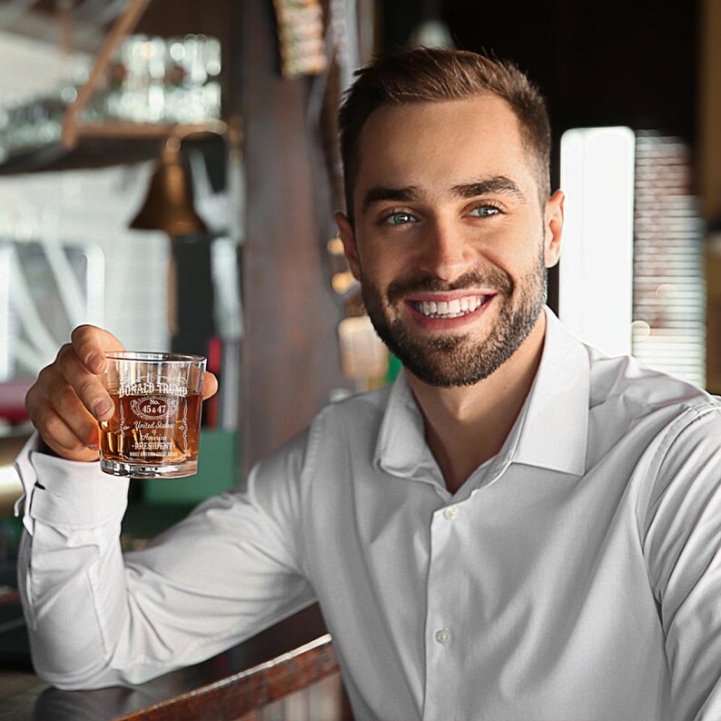 Peut inclure: Un homme portant une chemise blanche boutonn&eacute;e sourit en tenant un verre transparent contenant un liquide brun. Le verre porte une &eacute;tiquette indiquant "Jack Daniel's Tennessee Whiskey" et "Old No. 7 Brand".