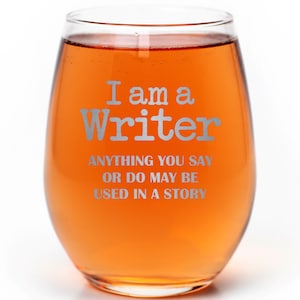 May include: A clear glass wine glass with a stem and a rounded base. The glass is filled with a reddish-orange liquid. The glass has the text "I am a Writer ANYTHING YOU SAY OR DO MAY BE USED IN A STORY" etched on the side.